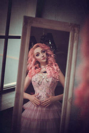 photo session of the girl porcelain doll girl with pink hair looking in the mirror and sees his reflection, baby doll, fashion creative color toningの写真素材