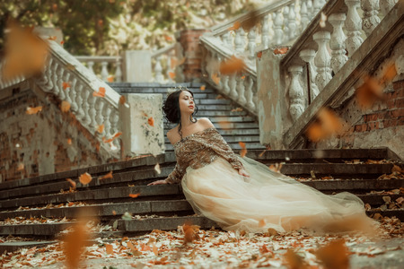 Beautiful girl in a luxurious dress walks in autumn park near the castle.Princess sits on the steps of the fall of the castle.Fashionable toning.creative colorsの写真素材