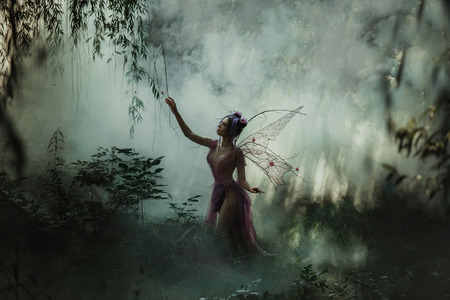 Fantastic photo shoot with smoke. Fairy Coquette walking in the fog. Background fairy forest, vines and trees. Unusual fabulous looks, purple dress and torn wings. Fancy toning.の写真素材