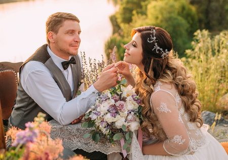 Sensual and luxurious Bridal couple on the shore of a picturesque lake.The groom lovingly looks at his bride and gently holds her hand.Retro style.Provence.Wedding bouquet, flowers.Fashionable toningの写真素材