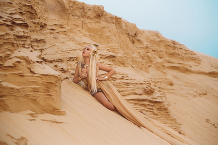Beautiful queen of the desert sitting on the sand. Luxury golden dress and unusual jewelry. Gold flower in her hair, hairstyle with braids. Beautiful blonde. Fashionable toning.の写真素材