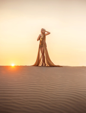 Beautiful queen of the desert at sunset walks. Luxury golden dress and unusual jewelry. Gold flower in her hair, hairstyle with braids. Beautiful blonde. Fashionable toning.の写真素材