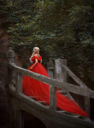 Beautiful blonde girl in a luxurious red dress. Queen goes on a beautiful staircase. Background Gothic wall with vines. Fairy Photo. Fashionable toning.の写真素材