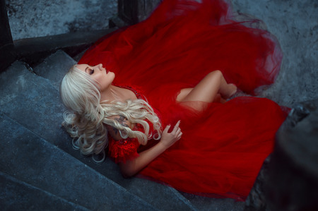 Beautiful blonde girl with long hair. She is in a luxurious red dress. The Queen on the beautiful staircase near her castle. Smoky eyes and red lips. Fairy Photo. Fashionable toning.の写真素材