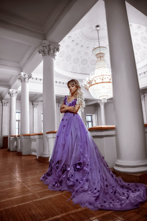 Beautiful, young, blond woman in a luxurious, purple, dress with a long train. The Queen walks into her castle. Fairy Photo. Creative toning. Background ballroom with a large chandelier.の写真素材