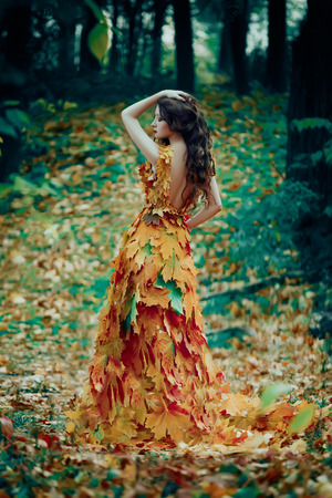 Fantastic girl in the autumn forest. Sad Queen of Nature in a luxurious dress with a leaf. Long hair. Creative colorsの写真素材