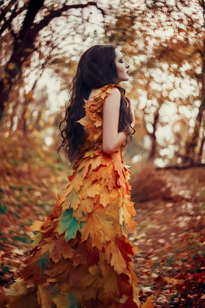Fantastic girl in the autumn forest. Sad Queen of Nature in a luxurious dress with a leaf. Long hair. Creative colorsの写真素材