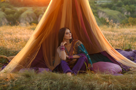 Beautiful woman with dark hair Oriental appearance in national costume sits in a unique fairy-tale hut. The image on the backdrop of the beautiful wildlife in the rays of the Golden sunset.Fashionable tinted.Creative color.の写真素材