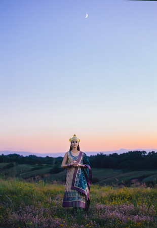 Beautiful girl with dark hair Oriental appearance, standing among fields of flowers, holding a candle in front of him.Image on the backdrop of scenic wildlife.Incredibly beautiful sky.Fashionable tinted.Creative color.の写真素材