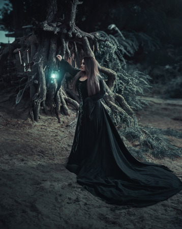 Beautiful large witch Halloween wearing vintage Gothic dress with long sleeves and sweeping train. Conjures in the dark night forest in the background, creepy tree. Fabulous and mystical concept. Fantastic shooting in the wild. Fashionable toning. Creativの写真素材