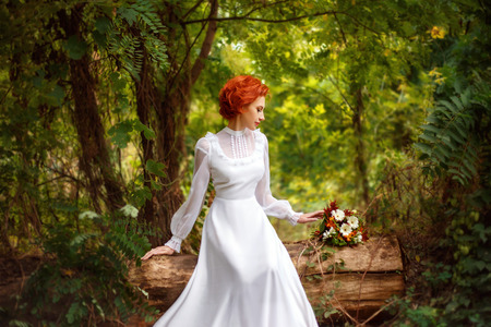 Gorgeous redheaded girl in white long dress sitting under the open sky and looking at the flowers....