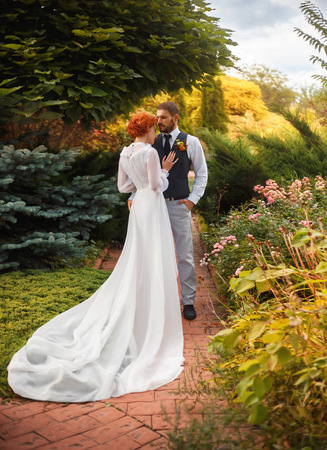 The young bridegroom looks tenderly at his beautiful bride. A girl stands leaning against to him in a beautiful garden. Dress long luxurious train. The air is filled with love. The picturesque nature.Fashionable toning. Creative color.の写真素材