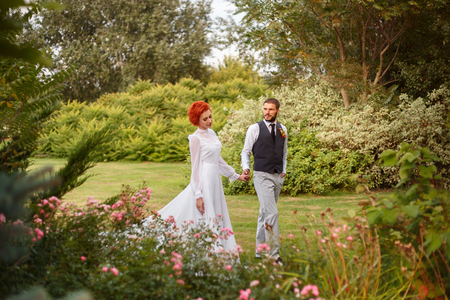The young bridegroom looks tenderly at his beautiful bride. The girl goes with him, hand in hand, in a beautiful garden. The air is filled with love. The picturesque nature.  Fantastic shooting. Retro style. Fashionable toning. Creative color.の写真素材