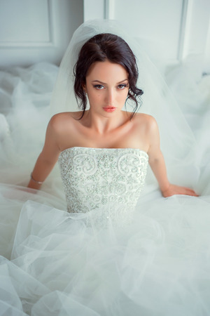Beautiful young girl sitting in a bright room.She is dressed in luxurious, fluffy, white dress.She looks in camera.European image of bride.Photo high key style.Fashionable toning.Creative color.の写真素材