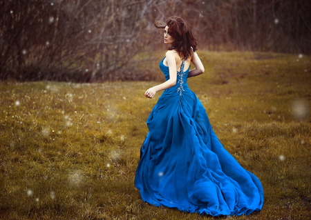 Beautiful brunette in blue lush dress with unusual back,she going forest trail looking back.Wind develops hair.Scenic background, branches of trees.Fantastic shooting.Fashionable toning.Creative colorの写真素材