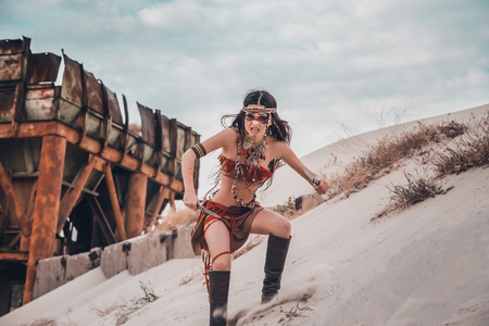 Warrior. Wild american indian girl ran to attack its prey. Ethnic shot against the background of an old rusty structure and sand. Unusual savage costume, jewelry, makeup combat. Fashionable toning.の写真素材