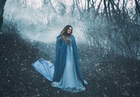 A big, beautiful woman in a blue raincoat, walks in a fog. Background dark forest, bare trees. Creative colorsの写真素材