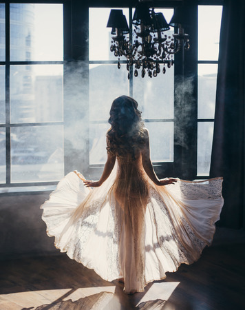The girl stands with her back to the camera on the background of a huge window. Transparent dress flies to the sides.の写真素材