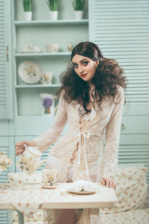 Girl with huge blue eyes posing on camera. The lady is pouring tea, in the old-fashioned kitchen. Creative hairstyle with a strong fleece. Make-up of the color of Marsala.の写真素材