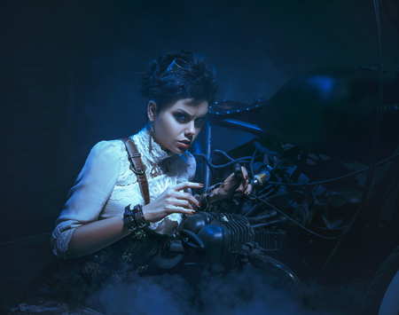 A beautiful, sexy girl is repairing a bike. Clothes in the steampunk style. Dark background with smoke. Creative colors.の写真素材