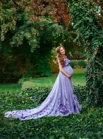 An elegant, pregnant woman walks in a beautiful garden in a luxurious, floral purple dress with a long train. Artistic Photographyの写真素材