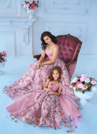 Mom and daughter in luxurious, pink dresses with sakura flowers on a skirt. Family clothes, identical dresses. The background is beautiful, expensive, white, classic interior. Artistic, family photoの写真素材