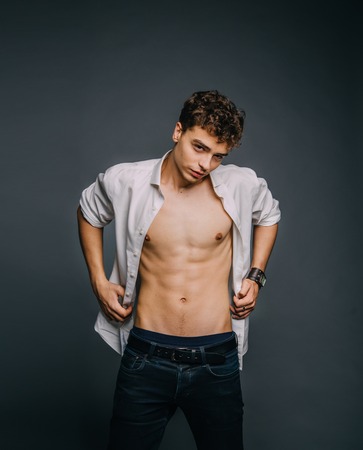 A young, handsome guy posing with an unbuttoned, white shirt, showing a sporty body, muscles, relief, pumped-up body. Background gray. Studio shooting, minimal, natural retouchingの写真素材