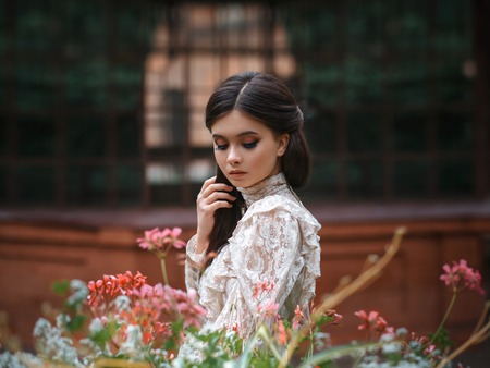 A girl walks in a flowering garden, she has a vintage blouse with a bow, chestnut long hair. she gently cares for her flowers. Sweetheart gardener. Artistic photography. Love of plantsの写真素材