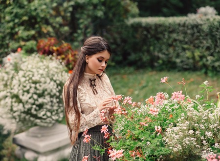 A girl walks in a flowering garden, she has a vintage blouse with a bow, chestnut long hair. she gently cares for her flowers. Sweetheart gardener. Artistic photography. Love of plantsの写真素材
