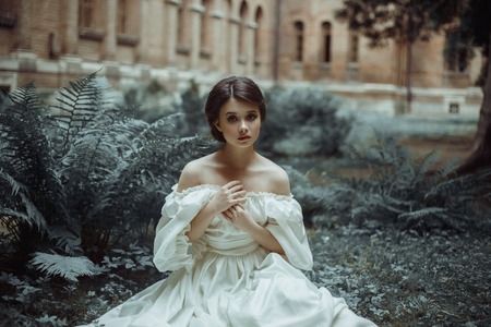 An incredibly beautiful princess sits in the castle garden amid the fern and moss. A beautiful, frightened face. Big sad eyes. Collected hair - a neat hair. On the lady - an old ivory dress. Art photoの写真素材