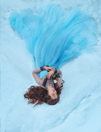 A sleeping beauty lies on the snow with a white rose in her hands. She is dressed in a luxurious, lush, blue dress. Light-haired, wavy, long hair is scattered in the snow. Artistic photoの写真素材