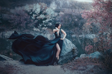 The dark queen pose against the background of gloomy rocks. A luxurious black dress with a long train fluttering in the wind denuding her leg. Elegant, collected hairstyle with a gothic crown.の写真素材