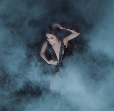 The dark vampire queen lies in the embrace of the fogs on her black dress with a deep neckline. The girl is enveloped in clouds of smoke, like blankets the clouds. Gothic atmosphere. Sleeping Beautyの写真素材