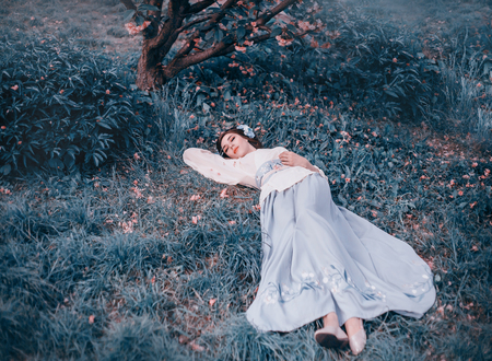 A young girl in a Japanese national kimono suit lies under a blooming sakura in pink petals. A Korean sleeping beauty. Artistic photography in cold shadesの写真素材
