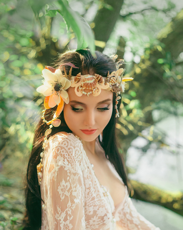 Close-up portrait - makeup of a river nymph, in gentle green, yellow, peach hues. The brunette girl with an unusual wreath with seashells. Puppets baby face. Wild nature background.の写真素材