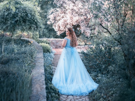 A big, beautiful, young woman in a luxurious blue dress with a long train is walking in a blooming garden. The model size is plus. The hair is large with curls. Artistic photoの写真素材