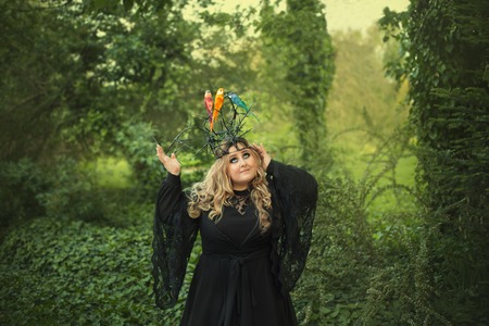 A big, beautiful, young woman in a black dress and in a crown with thorns on which birds are sitting-wavy parrots. The Queen walks in the garden against the background of trees overgrown with ivyの写真素材
