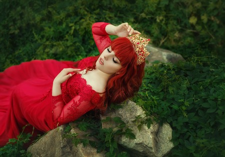 The Queen in a luxurious, expensive, red dress, with a long train lies on the thickets of ivy. Red-haired girl in a gold crown. The background is cold, juicy greens. Art photoの写真素材