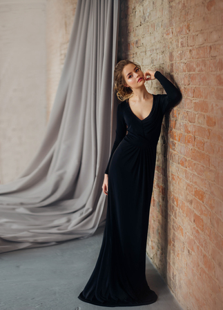 A young blonde girl in a strict, discreet, modest, elegant, long black dress. Delicate natural make-up. Beautiful, elegant, high hairstyle for an average length of hair, with weaving. Evening imageの写真素材