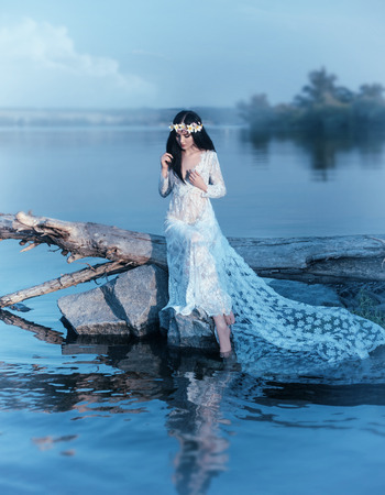 A nymph with long dark hair in a white vintage dress sits on a log across the river. in the hair a wreath of lilies. Fantasy photosession, artistic processing in cold blue tonesの写真素材