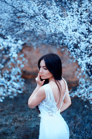 wonderful attractive lady with dark black long hair and blue eyes, standing in an arch of blooming magnolias, dressing luxurious, gorgeous white snow-white wedding dress with an open back, cold colorsの写真素材