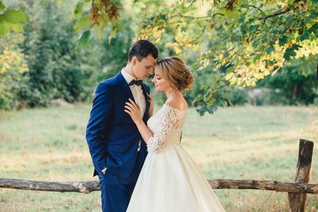 Amazing attractive young couple with closed eyes in wedding day. bride in elegant white long dress and blue bouquet in hand, the groom in a blue fashionable business suit, standing near the lawnの写真素材