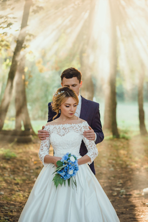 Amazing attractive young couple in wedding day. bride in elegant white long dress and blue bouquet in hand, groom in a blue fashionable business suit in soft sun rays breaking through trees leavesの写真素材