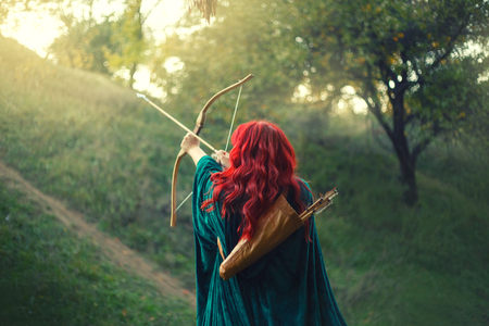 gorgeous huntress pushing her last light to the sun, waiting for salvation during terrible danger, red-haired girl fights bravely for life, emerald velvet cloak, photo without a face from the backの写真素材