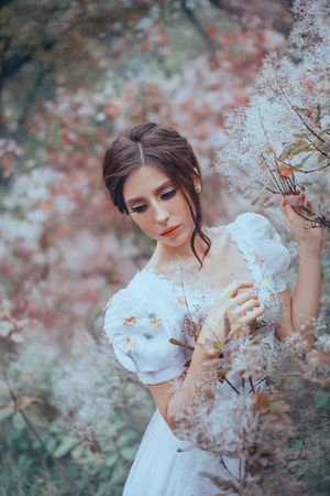 mysterious lady in an expensive light vintage dress with patterns stands by flowering trees, one with a sad look, lowered her eyes, dark hair gathered in a gentle hairstyle, curls fall on her faceの写真素材