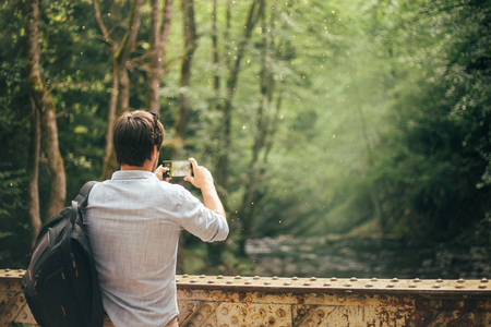 experienced fashionable dark-haired tourist, man takes a photo on his new phone, advertising and announcement, the delightful nature of Georgia, bright green and emerald tree colorsの写真素材