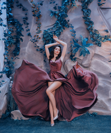 slender attractive lady like picture of gorgeous artist, flying fluttering marsala long satin dress like paint strokes, sexy girl elegantly raised her arms top, shows charming bare legs in gray heelsの写真素材