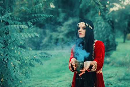 wise fortune-teller in silk red scarlet dress and gold decoration stands alone in forest with magic bowl, calling spirits white and light smoke, Turkish beauty with passionate look poses to cameraの写真素材
