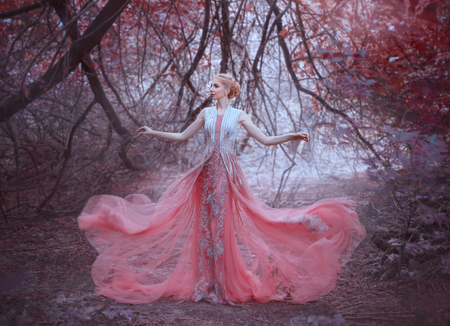 attractive girl with gorgeous blond hairdo in the forest near the branches of trees, dressed in a light amazing pink tender dress, throws it down, flying fluttering clothes, art photo, creative colorの写真素材