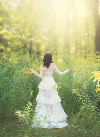 charming sweet girl with dark hair and bare shoulders stands in gorgeous white dress with her back to camera in forest, princess of spring in white tender light dress in rays of bright sunの写真素材